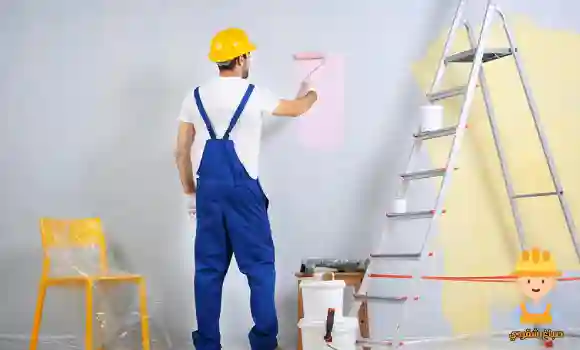 صباغ كبد يقوم بطلاء جدار باستخدام بكرّة الدهان مع تجهيزات العمل حوله في موقع داخلي